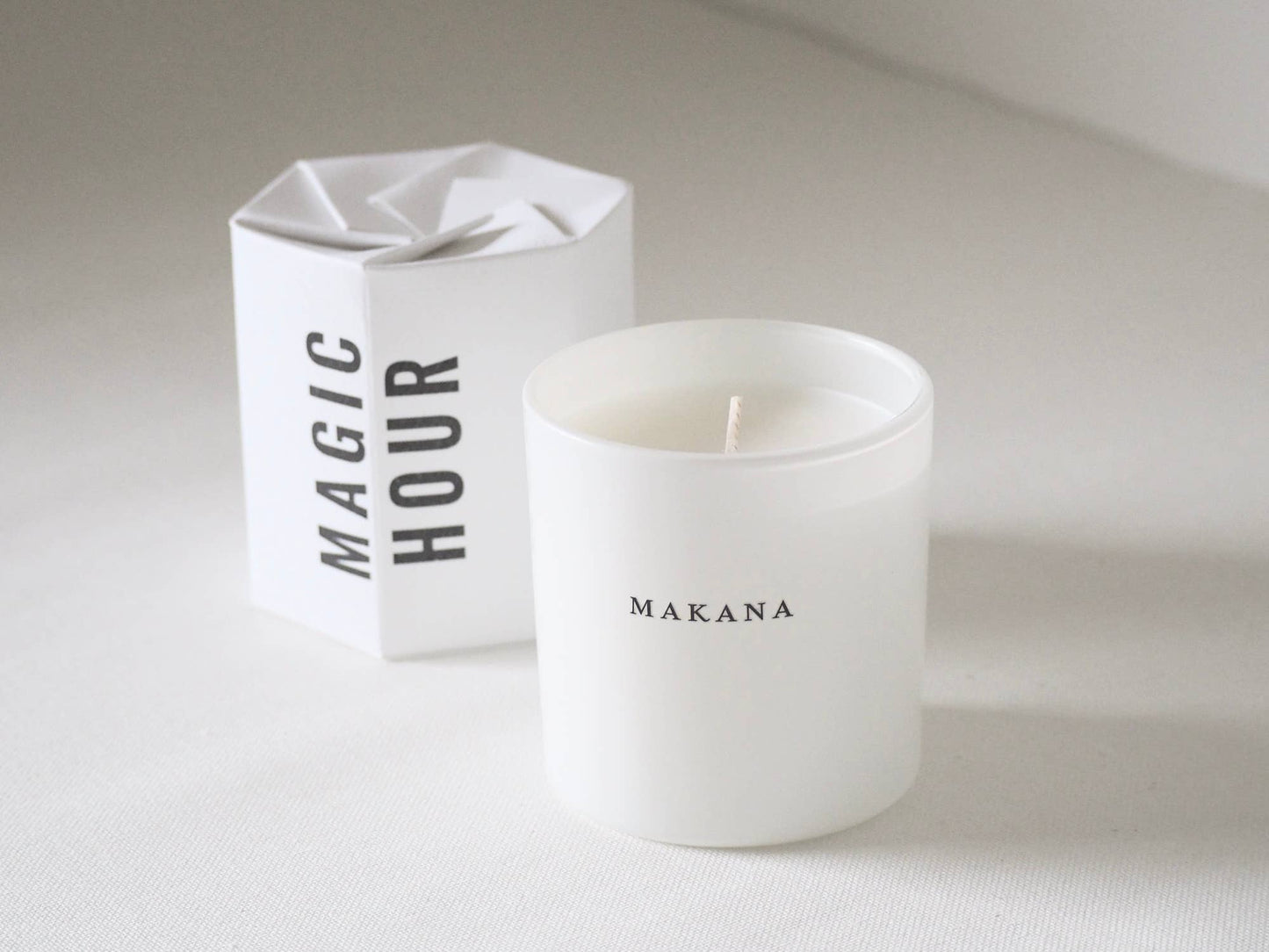 White candle with 'Makana' branding next to a white box labeled 'Magic Hour' on a light background
