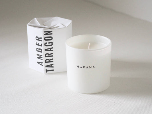 White candle with 'Makana' branding next to a white box labeled 'Amber Tarragon'.