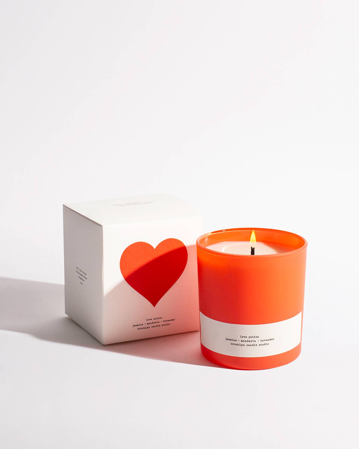 A red glass candle with a minimalist cream-textured label, sitting next to its white box with a heart-shaped cut-out lid.