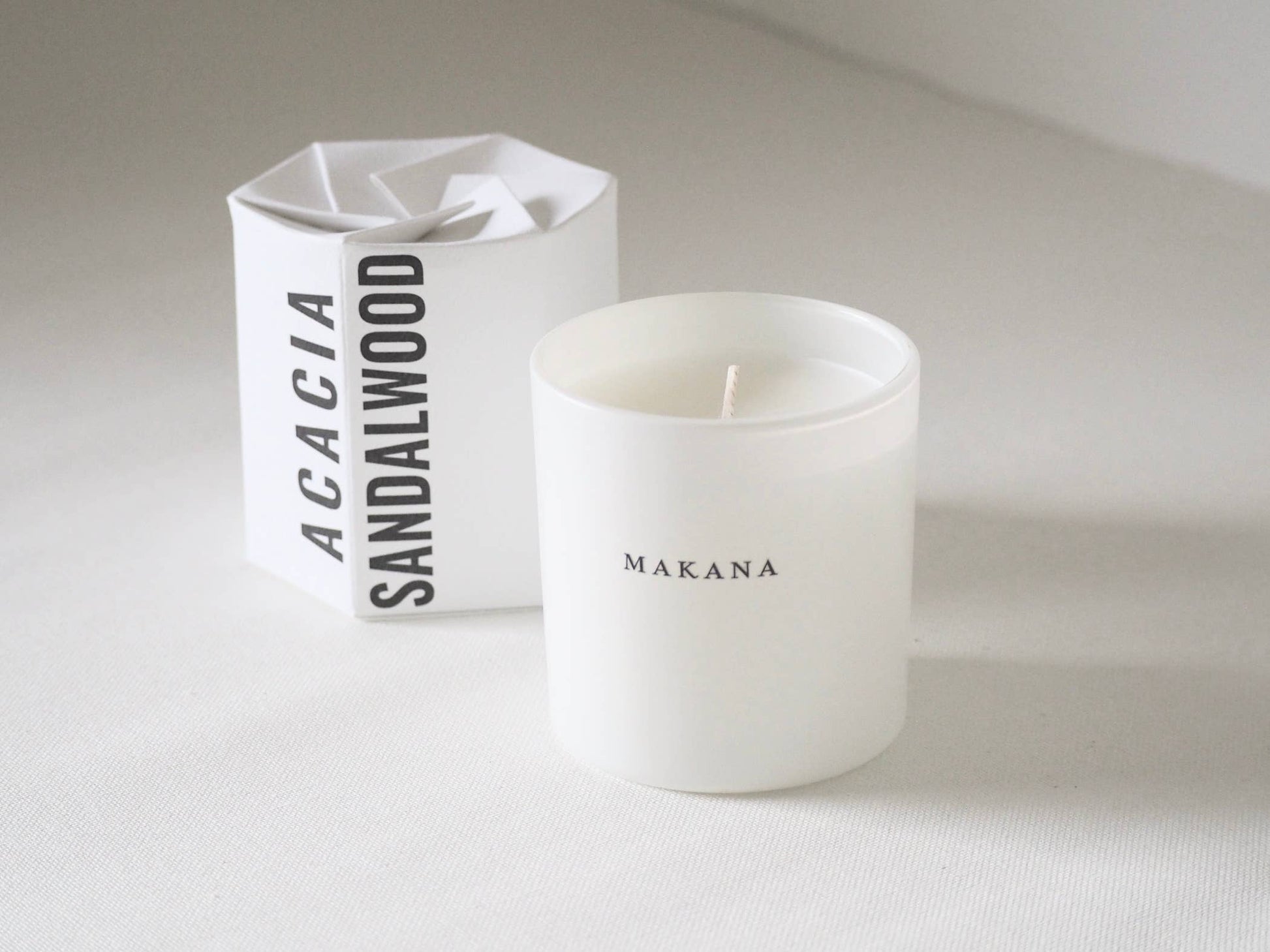 White candle with 'Makana' branding next to a white box labeled 'Acacia Sandalwood'.