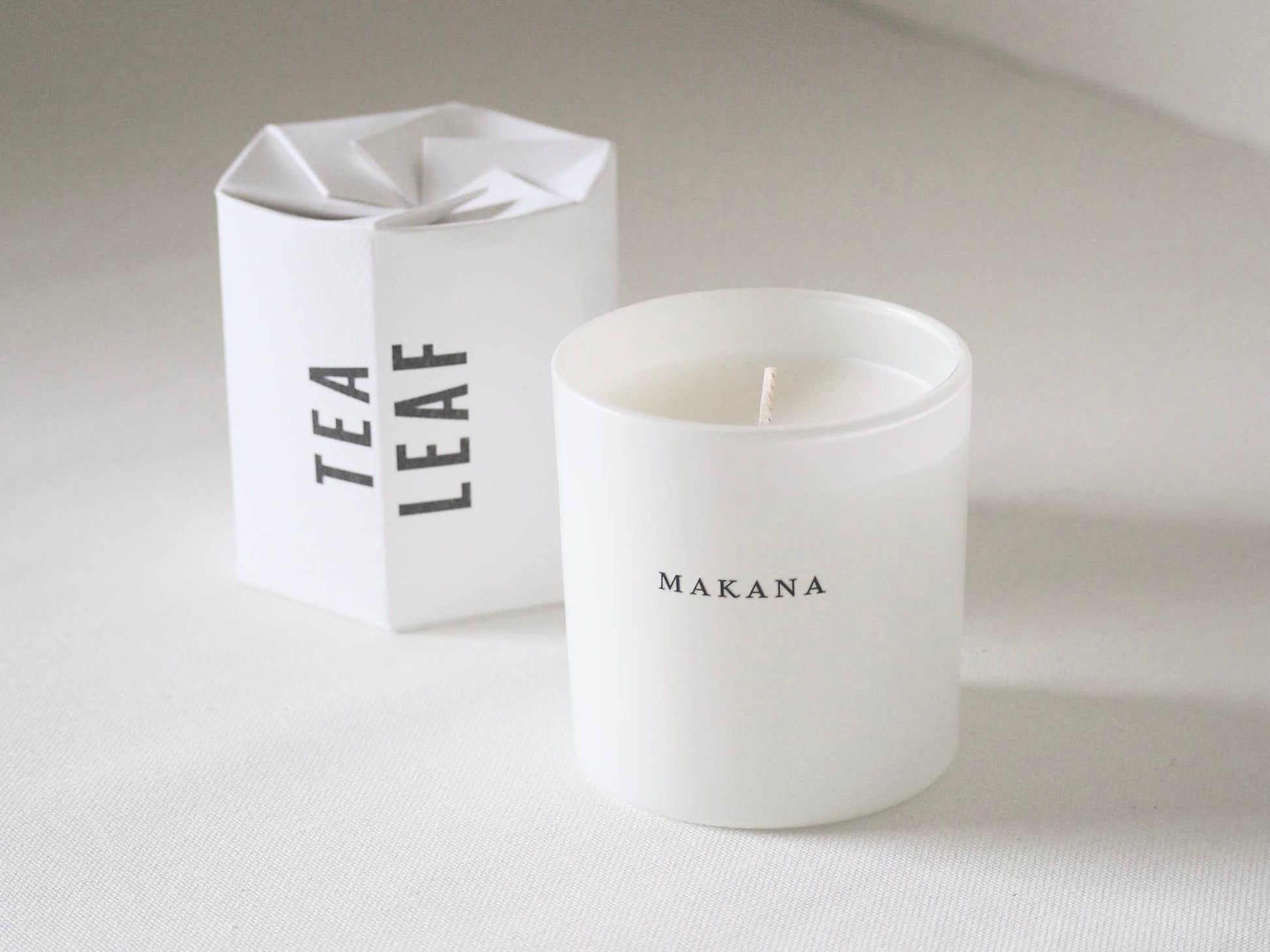 White candle with 'MAKANA' branding next to a white box labeled 'TEA LEAF' on a light background