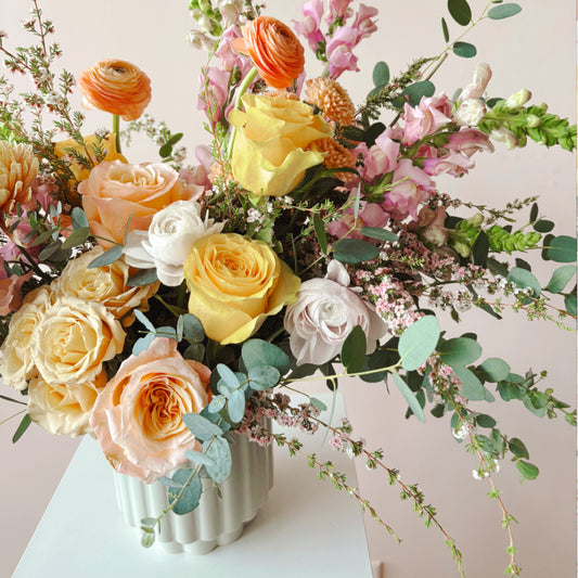 a close up image of yellow roses, peach ranunclus in a cream vase.