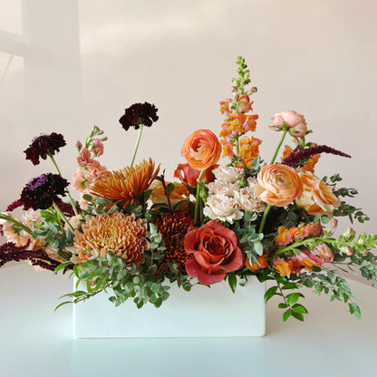A low and long thanksgiving flower arrangement in a rectangular vase with roses, ranunculus, snapdragons and mums