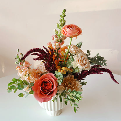 I beautiful, fall colors Thanksgiving flower arrangement featuring roses, ranunculus and stock in a petite pedestal vase.